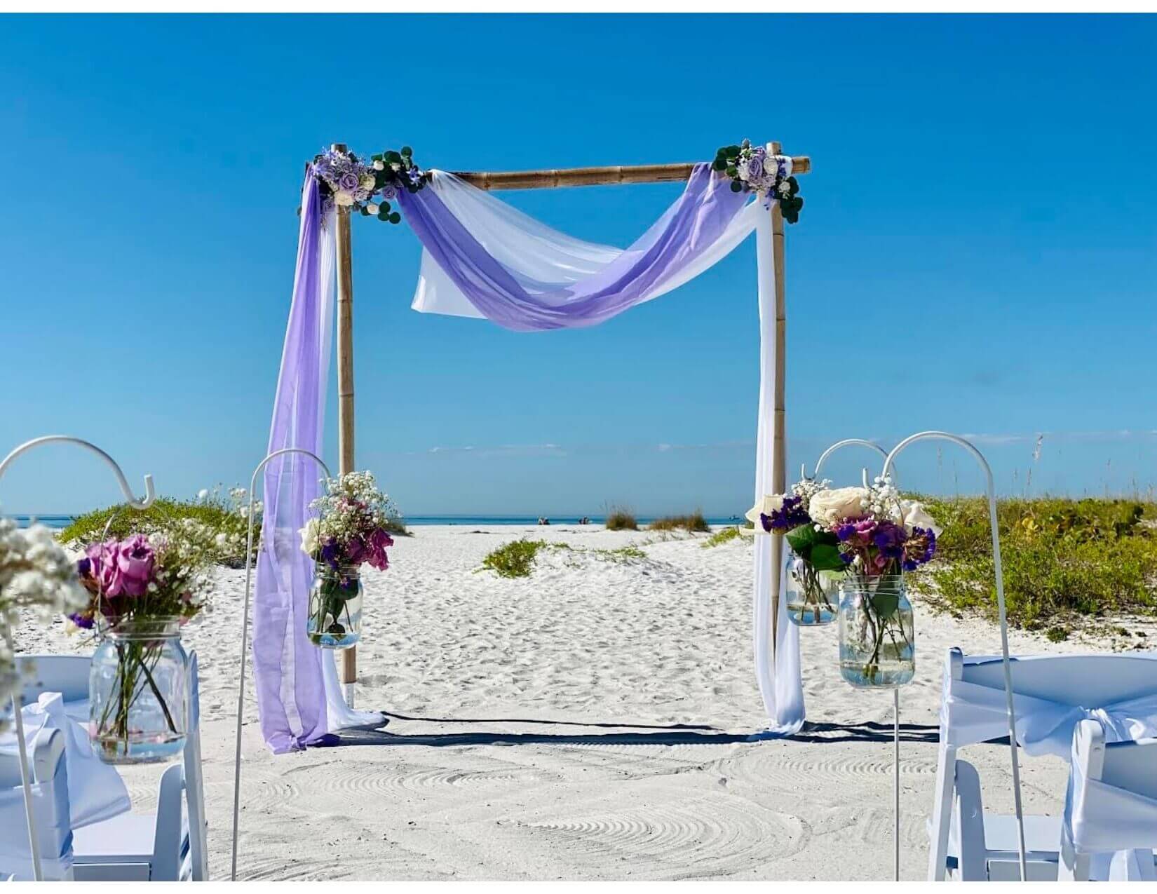 Bamboo two-post beach arch draped in lavender fabric with floral hanging jars on the sand — the Breath of Love package