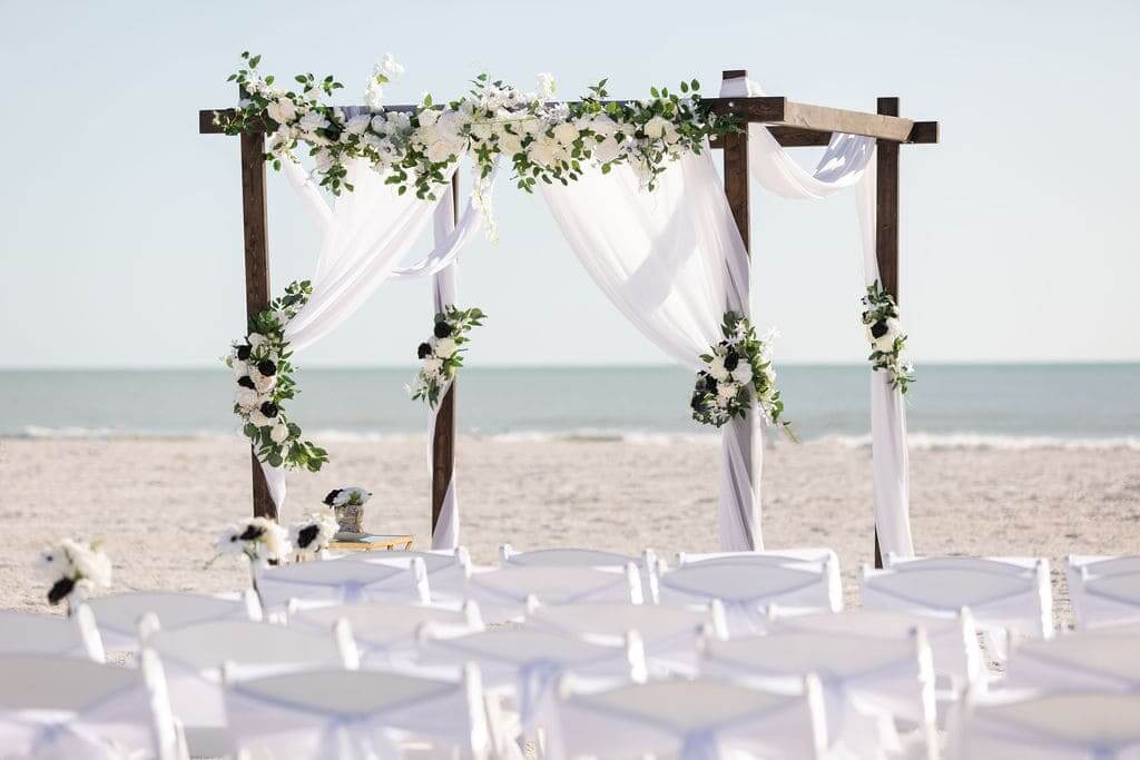 Wooden 4-post beach arch with white drapes and black-and-white florals over rows of white chairs — the Deep in Love package