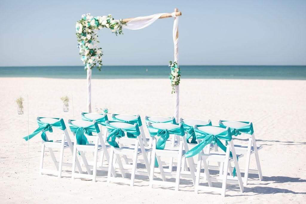 Beach ceremony aisle with white chairs, royal-blue sashes and a heart welcome sign — the Sunshine Kiss package