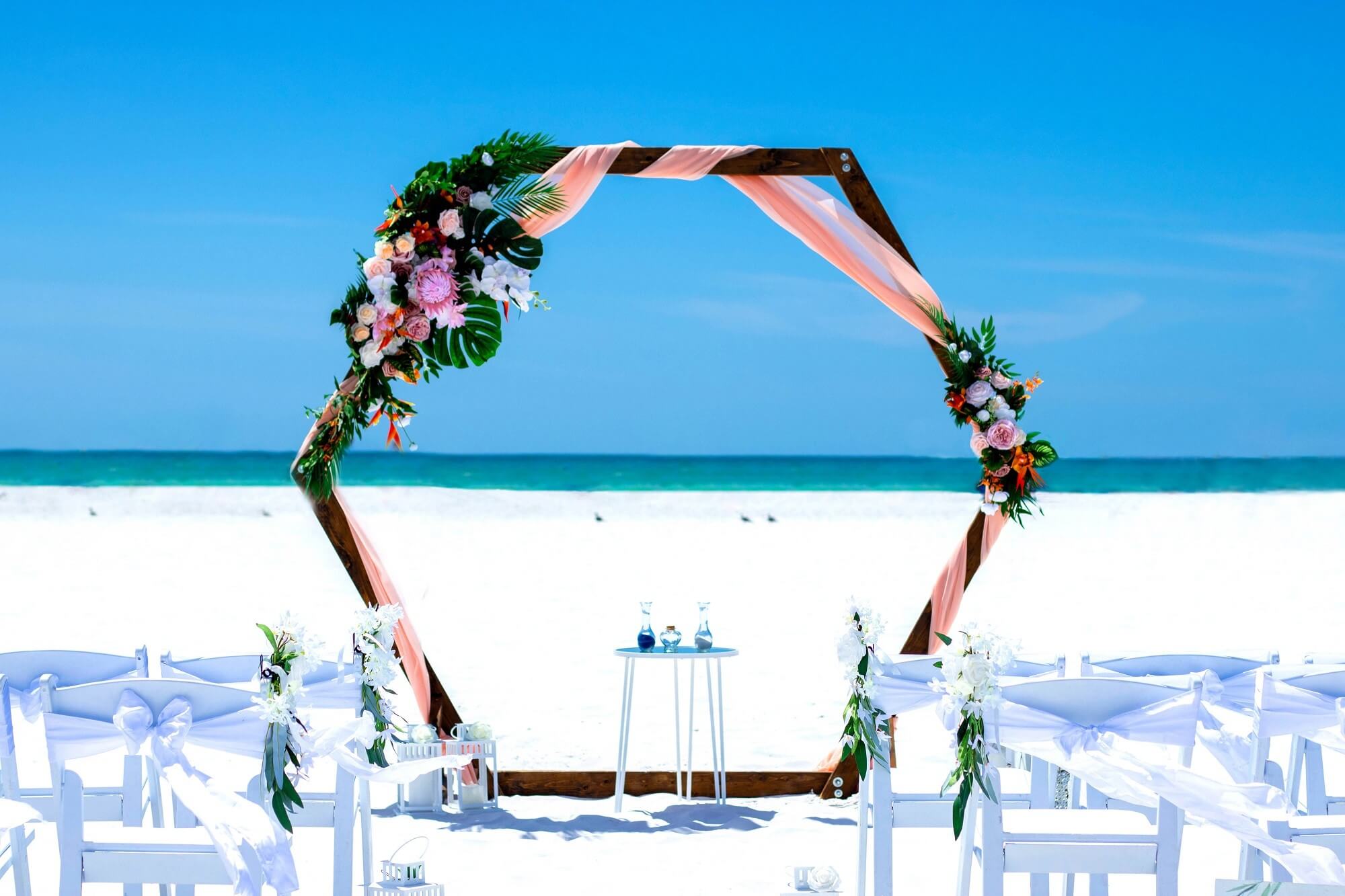 Wooden beach arch dressed with tropical monstera and palm leaves against a Gulf Coast sunset — the Tropical Bliss package